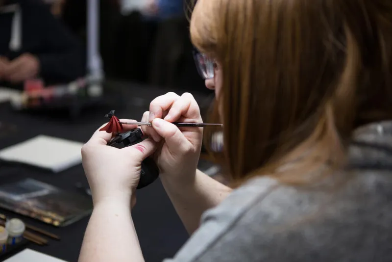 Woman with glasses carefully painting a small black and red miniature figure with a fine brush