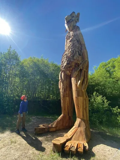 Person looking up at a towering wooden troll sculpture surrounded by green trees