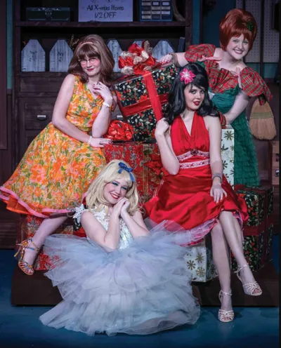 Four women in retro costumes pose among wrapped Christmas gifts on a theater stage