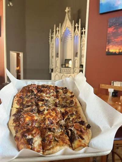 Square-cut flatbread pizza in parchment-lined tray with a decorative gothic-style altar piece in background
