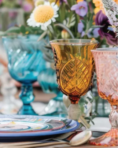 Colorful amber, teal, and pink embossed glass goblets on a decorated table with wildflowers
