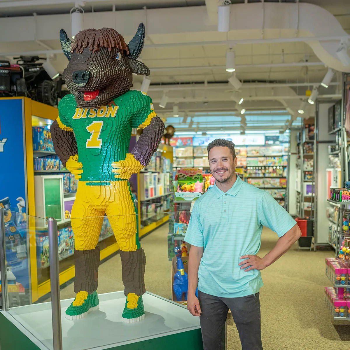 Smiling man in mint polo poses beside a life-size LEGO bison mascot in a green-and-yellow jersey inside a retail store