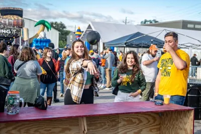 Two smiling women hold drinks at an outdoor festival crowd near tents and inflatable decorations
