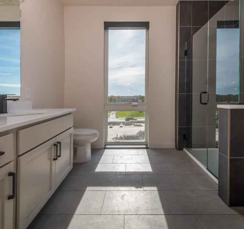 Modern bathroom with white vanity, gray tile floor, glass shower, and tall window overlooking a sunny landscape