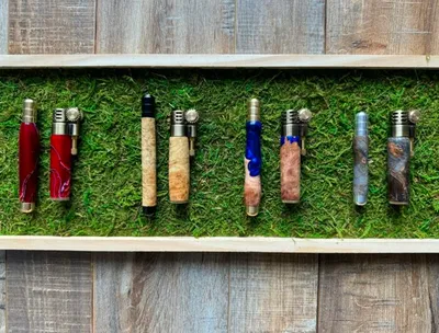 Eight handcrafted pens with colorful wood and resin bodies displayed on a mossy shelf