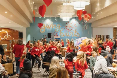 Crowd in red shirts gathered indoors beneath heart balloons near a 'Giving Hearts' wall mural