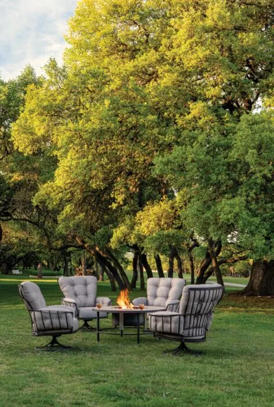 Four cushioned patio chairs arranged around a lit fire pit on a grassy lawn beneath large oak trees