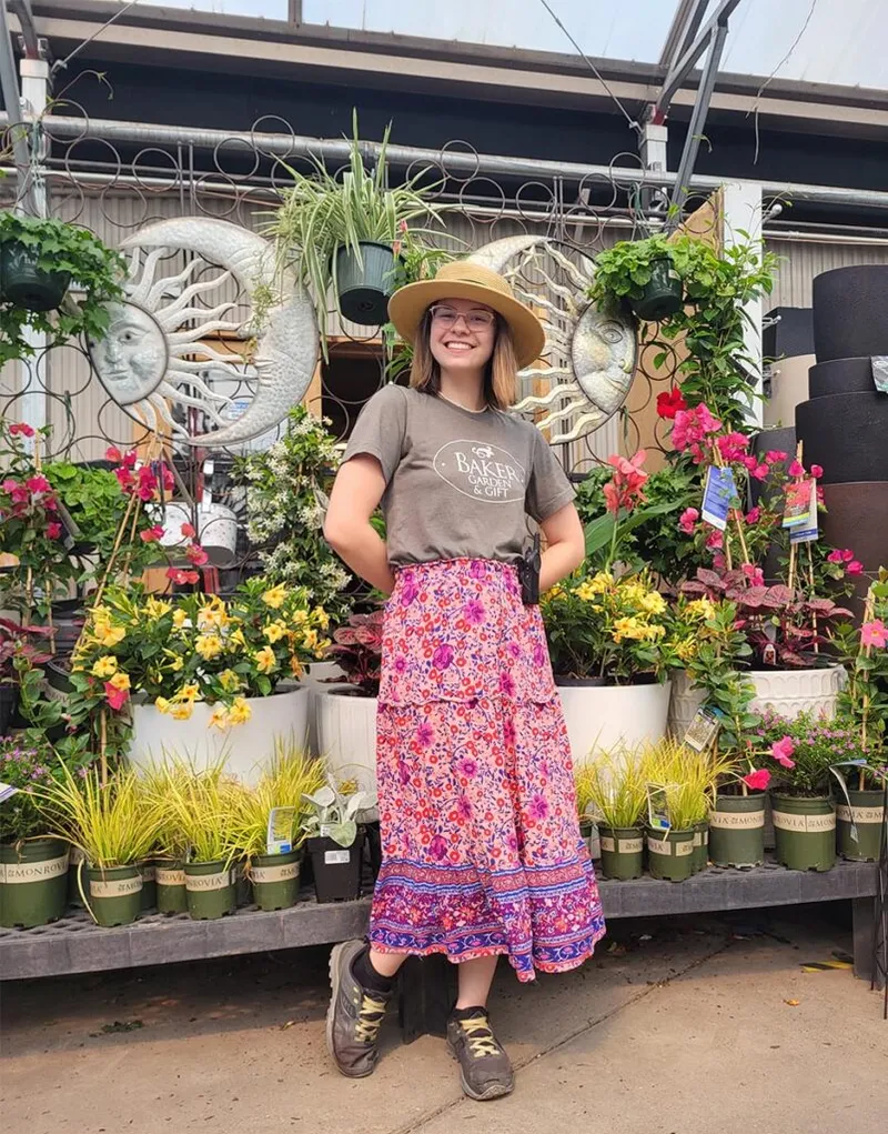 Smiling young woman in straw hat and floral skirt stands before colorful plants at a garden center