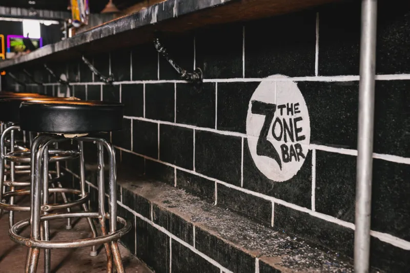 Black tiled bar interior with metal stools and a painted Zone Bar logo on the wall