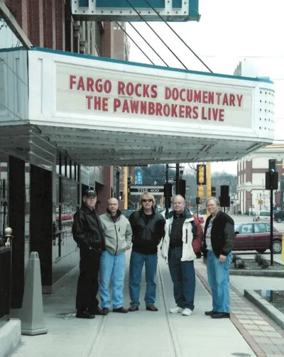 Five people standing beneath a theater marquee reading 'Fargo Rocks Documentary – The Pawnbrokers Live