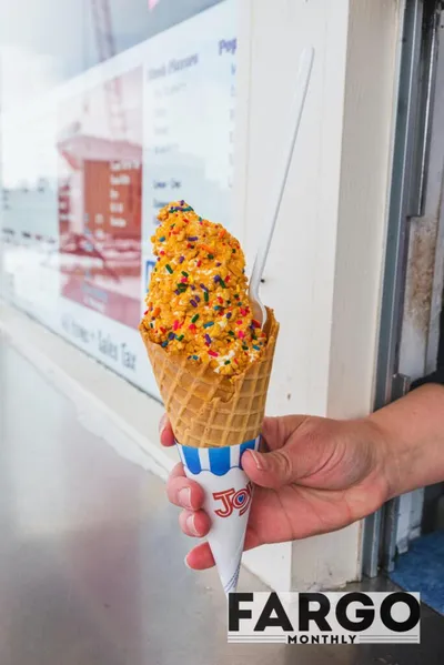 Hand holding a waffle cone topped with orange soft-serve and colorful sprinkles