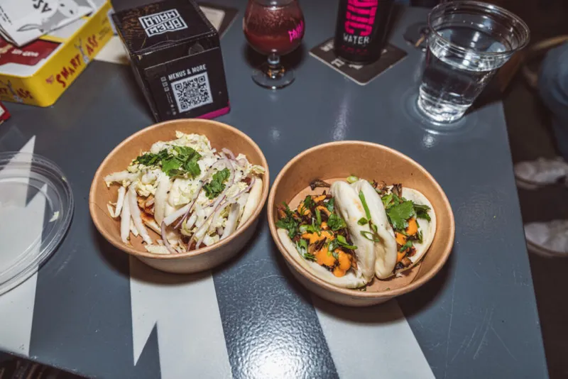 Two kraft bowls on a gray table — a slaw salad and steamed bao buns topped with cilantro and orange filling