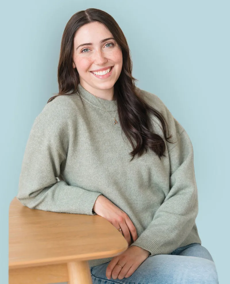 Smiling young woman with dark hair in a sage green sweater seated at a wooden table against a light blue background