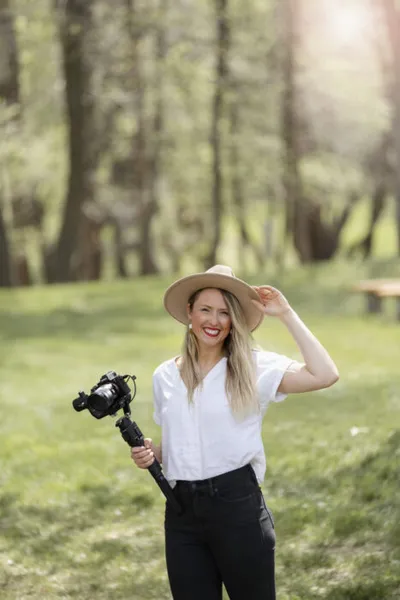 Smiling woman in wide-brim hat and white shirt holding a camera on a gimbal stabilizer outdoors