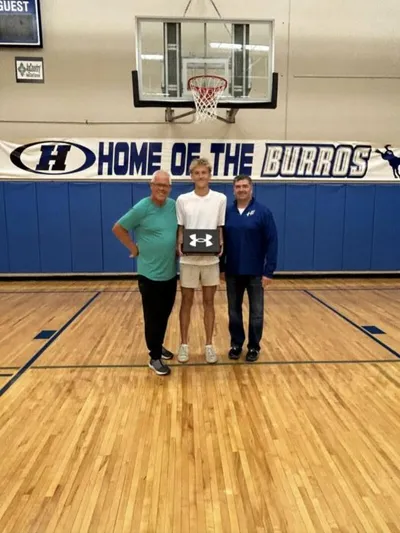Three people posing on a school gymnasium floor holding an Under Armour box, with a 'Home of the Burros' banner behind them