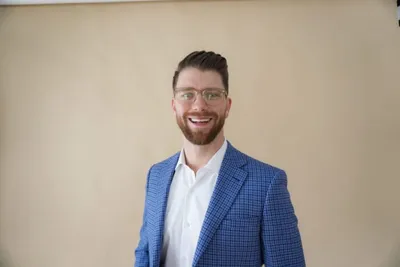 Smiling bearded man with glasses wearing a blue blazer and white shirt against a beige wall
