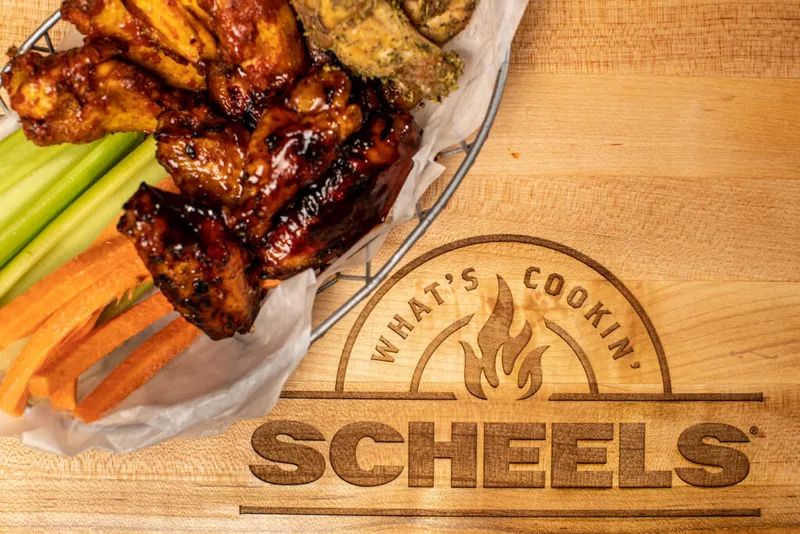 Glazed chicken wings with celery and carrots in a wire basket on a wood board engraved with the Scheels logo
