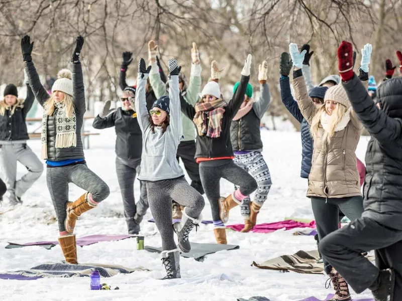 Group of people in winter coats and hats doing tree pose yoga on snow-covered ground