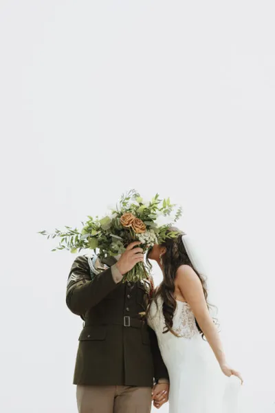 Couple kissing behind a lush green and amber rose bouquet, bride in white lace gown