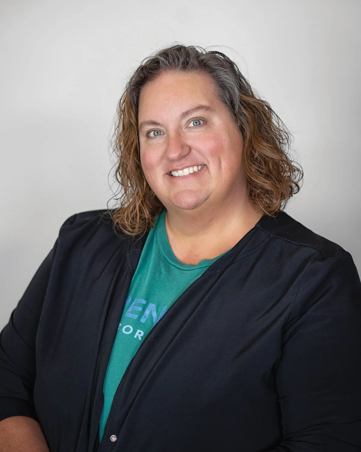 Smiling woman with curly brown hair wearing a teal shirt and black cardigan against a gray background