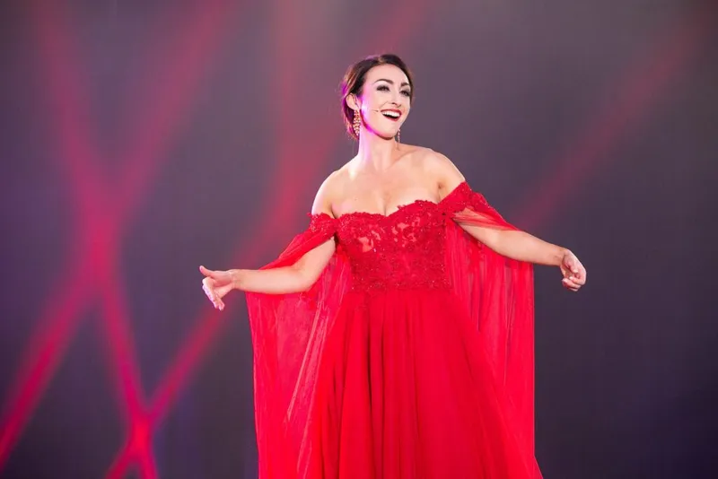 A woman in a flowing red off-shoulder gown performs on a stage under dramatic pink lights