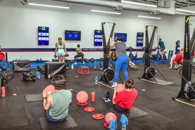 Group fitness class in a gym with people using weights, med balls, and cable machines on rubber flooring