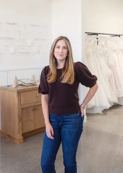 Woman in dark sweater and jeans standing in a bridal boutique with white gowns on racks behind her