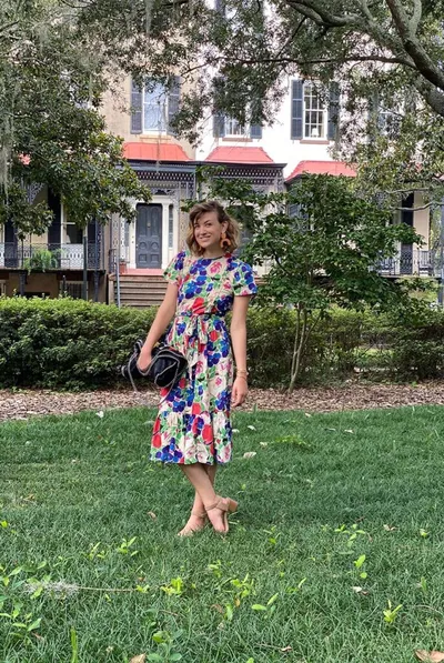 Woman in a floral midi dress holding a black handbag, standing on grass in front of a historic building