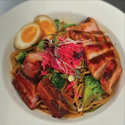 Bowl of noodles topped with glazed pork belly, broccoli, soft-boiled egg, and pink microgreens