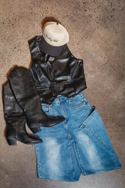 Flat lay of black leather vest, cowboy boots, denim shorts, and embroidered baseball cap on concrete