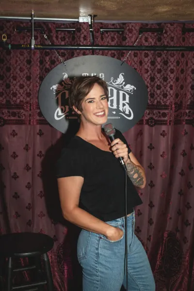 Tattooed woman in black tee and jeans holds a mic on a comedy club stage