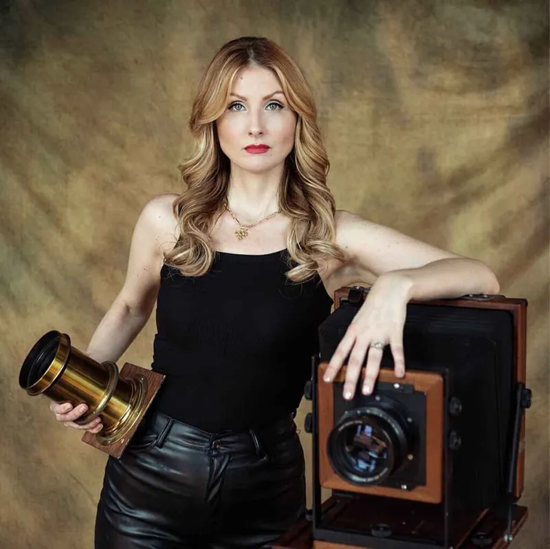 Woman in black tank top holding a brass lens beside a vintage large-format wood camera