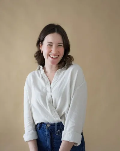 Smiling woman with dark hair in a white linen shirt and blue jeans against a beige backdrop