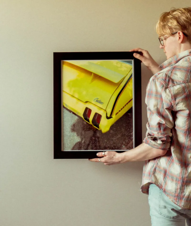 Person in plaid shirt hanging a framed photo of a yellow classic car on a wall