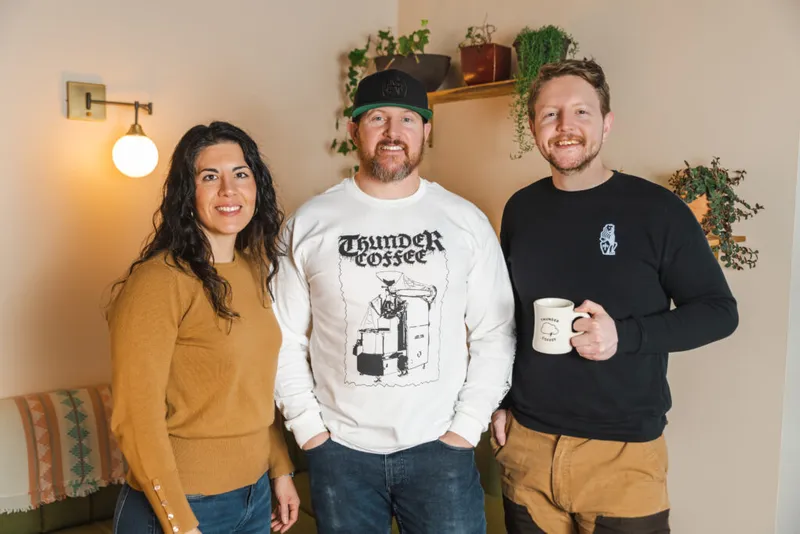 Three smiling people posing indoors, center wearing a Thunder Coffee sweatshirt, right holding a Thunder Coffee mug