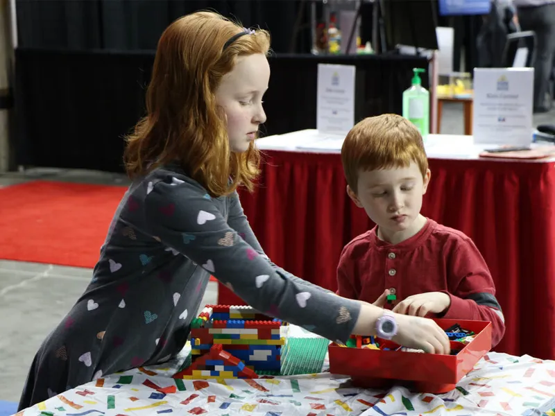 Two red-haired children building with colorful bricks at an indoor expo table