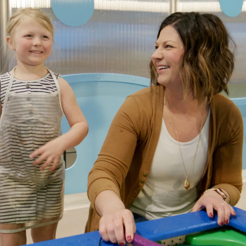 Smiling woman in brown cardigan with young blonde girl at a colorful play table indoors