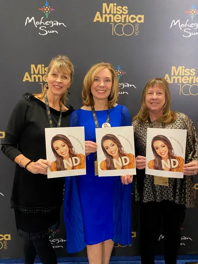 Three women pose in front of a Miss America 100 Years backdrop, holding Miss North Dakota programs