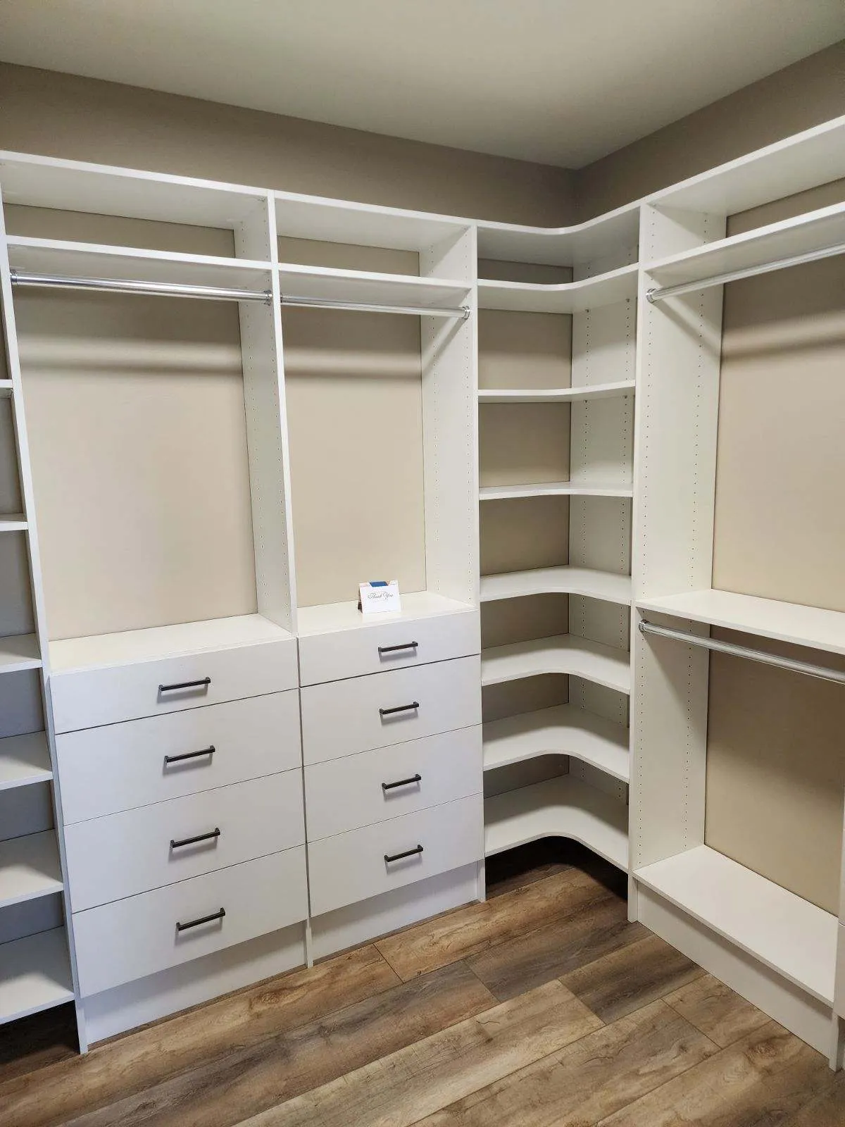 Empty walk-in closet with white built-in drawers, hanging rods, and corner shelving over wood-look flooring