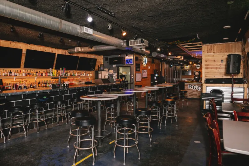 Empty bar interior with rows of black stools, a fully stocked back bar, and exposed ductwork overhead