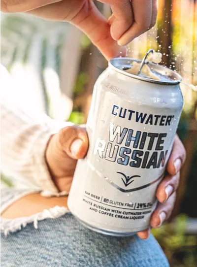 Hands opening a Cutwater White Russian canned cocktail with fizz spraying out