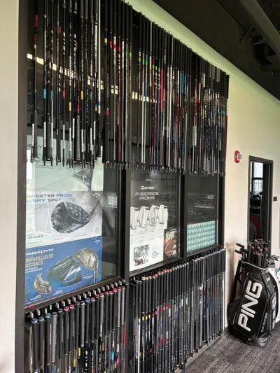 Wall display of golf club shafts with brand posters and a Ping golf bag to the right
