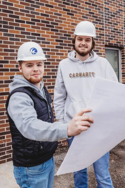 Two young men in white hard hats reviewing blueprints outside a brick building