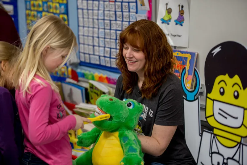 Smiling woman holds green alligator plush while a girl brushes its teeth in a colorful classroom