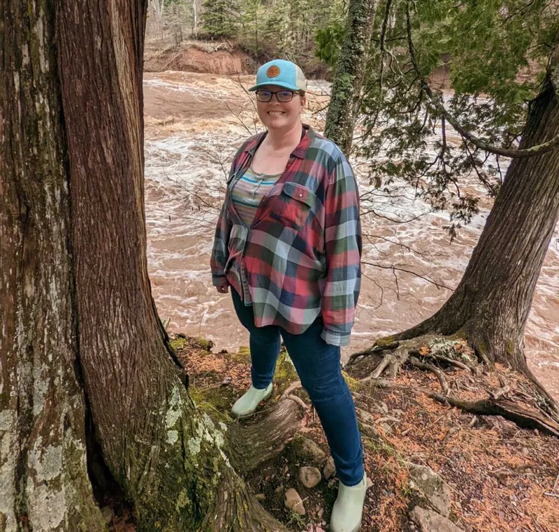 Smiling woman in plaid shirt and blue cap standing beside trees above a rushing muddy river