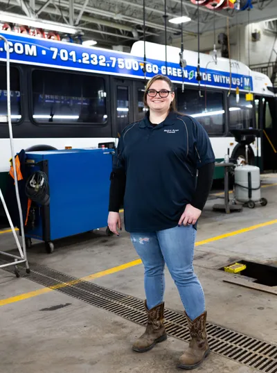 Woman in dark polo and jeans stands smiling in a bus maintenance garage