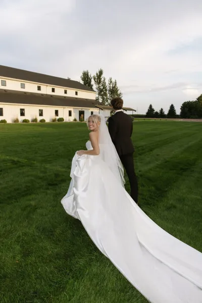 Bride glancing back over shoulder beside groom on green lawn outside a large white barn venue