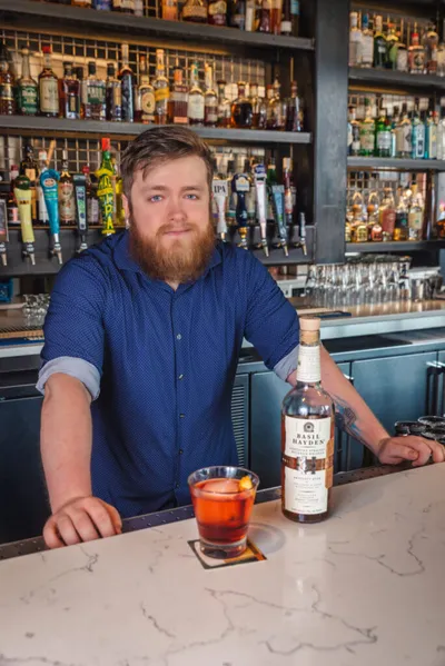 Bearded bartender in blue shirt leans on marble bar beside a cocktail and bourbon bottle