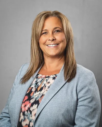 Smiling woman with blonde highlights wearing a light blue blazer and floral top against a gray backdrop