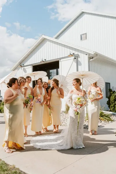 Bride in white lace gown with bridesmaids in yellow dresses holding paper parasols outside a white barn venue